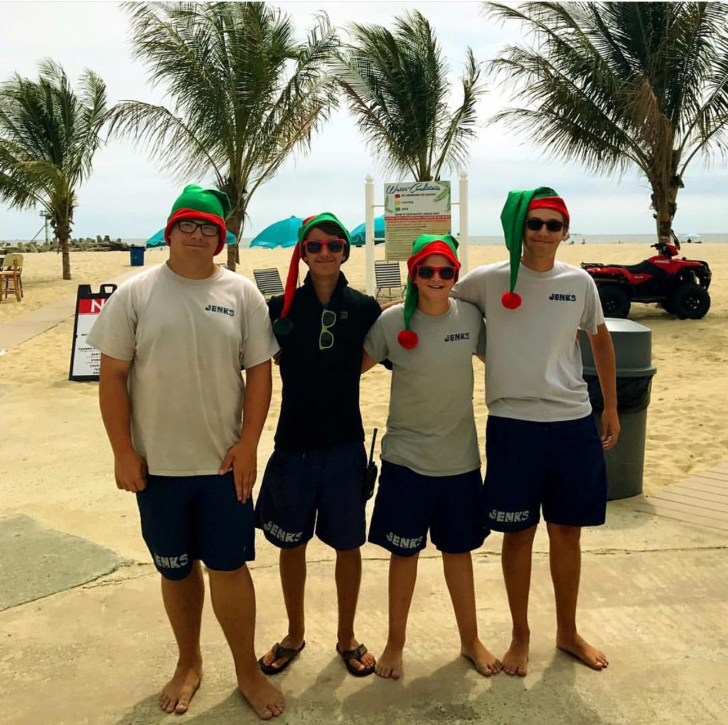 Four Jenkinson's employees lined up on the beach all wearing elf hats. Palm trees in background.