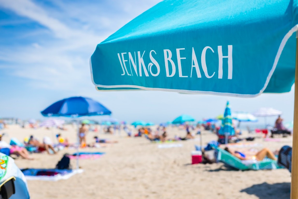 BEACH PHOTO A turquoise beach umbrella labeled "Jenks Beach" with people relaxing on the sandy beach in Point Pleasant Beach, NJ.