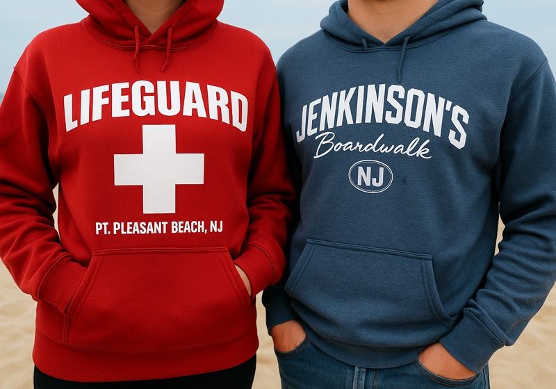 Two people wearing a red "Lifeguard" hoodie and a blue "Jenkinson's Boardwalk" hoodie standing on a sandy beach.