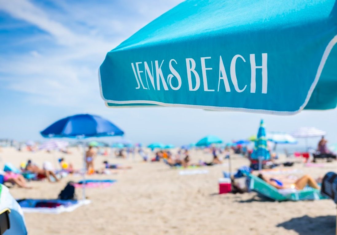 A turquoise beach umbrella labeled "Jenks Beach" with people relaxing on the sandy beach in Point Pleasant Beach, NJ.