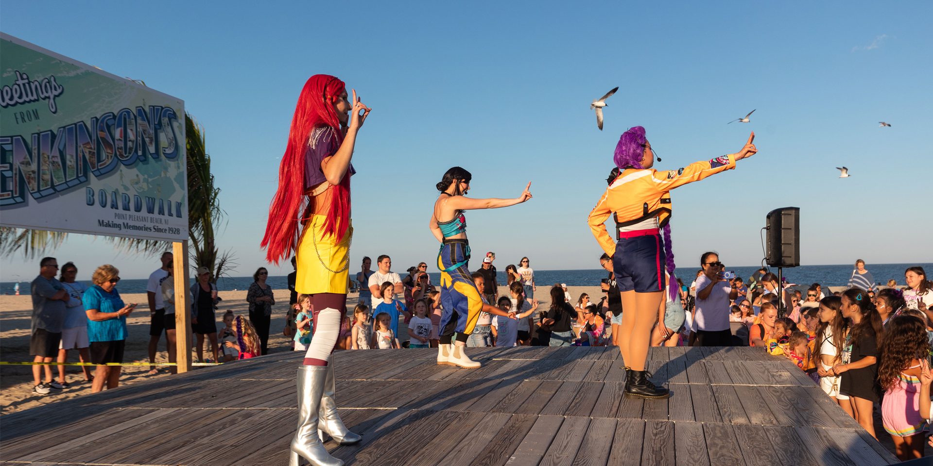 Performers in colorful costumes entertain a crowd of families during a live dance show on the South Beach Stage at Jenkinson's Boardwalk, with the beach and ocean in the background.